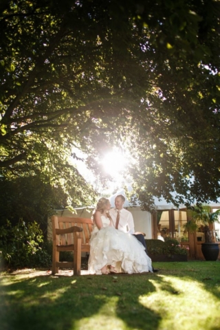 Bride and Groom sat on bench with sun shining between leaves