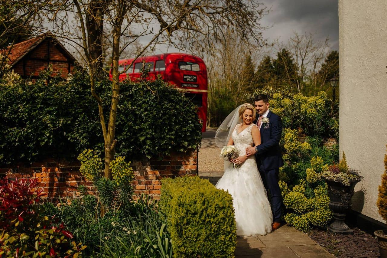 Couple standing on path by wall and red bus at wedding venue essex